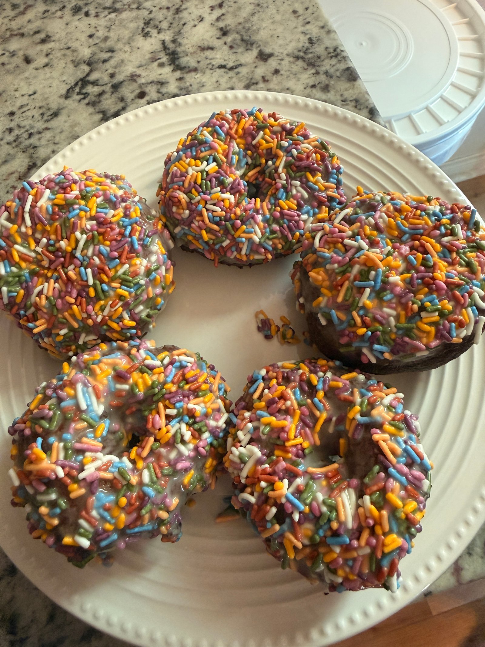 Five fresh milled doughnuts topped with white glaze and rainbow sprinkles on a white plate