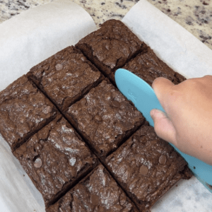 Cutting into brownies with a teal silicone bowl scraper. Brownies are mostly cut and shows brownies cut in a 3x3 grid into squares