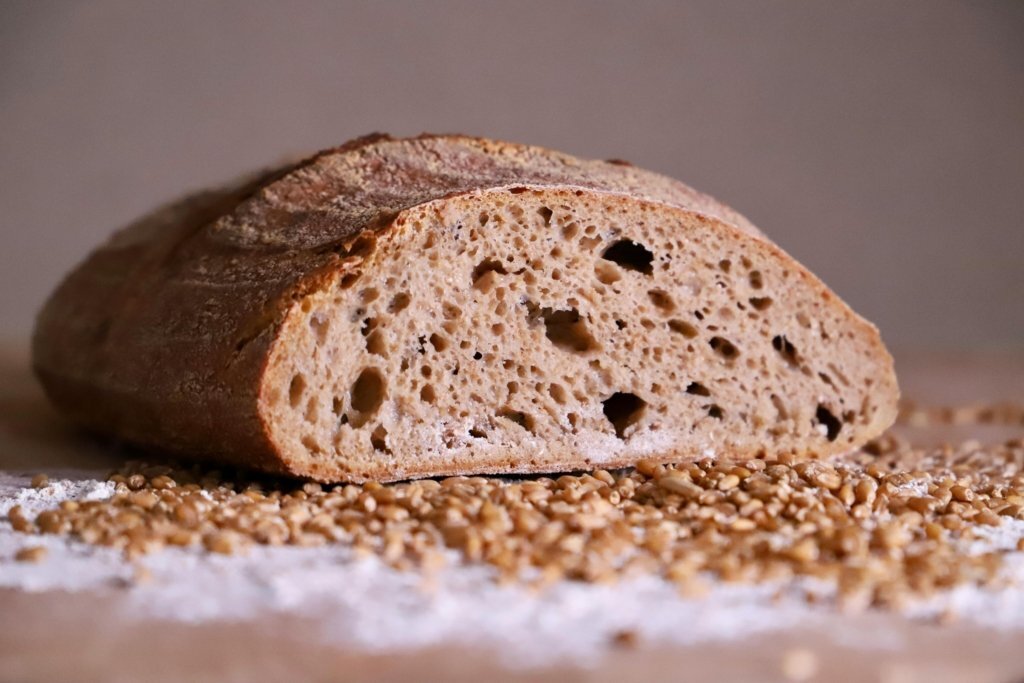 Close-up of a rustic artisan bread loaf slice surrounded by scattered wheat grains on a flour-dusted surface.