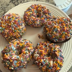 Five fresh milled doughnuts topped with white glaze and rainbow sprinkles on a white plate