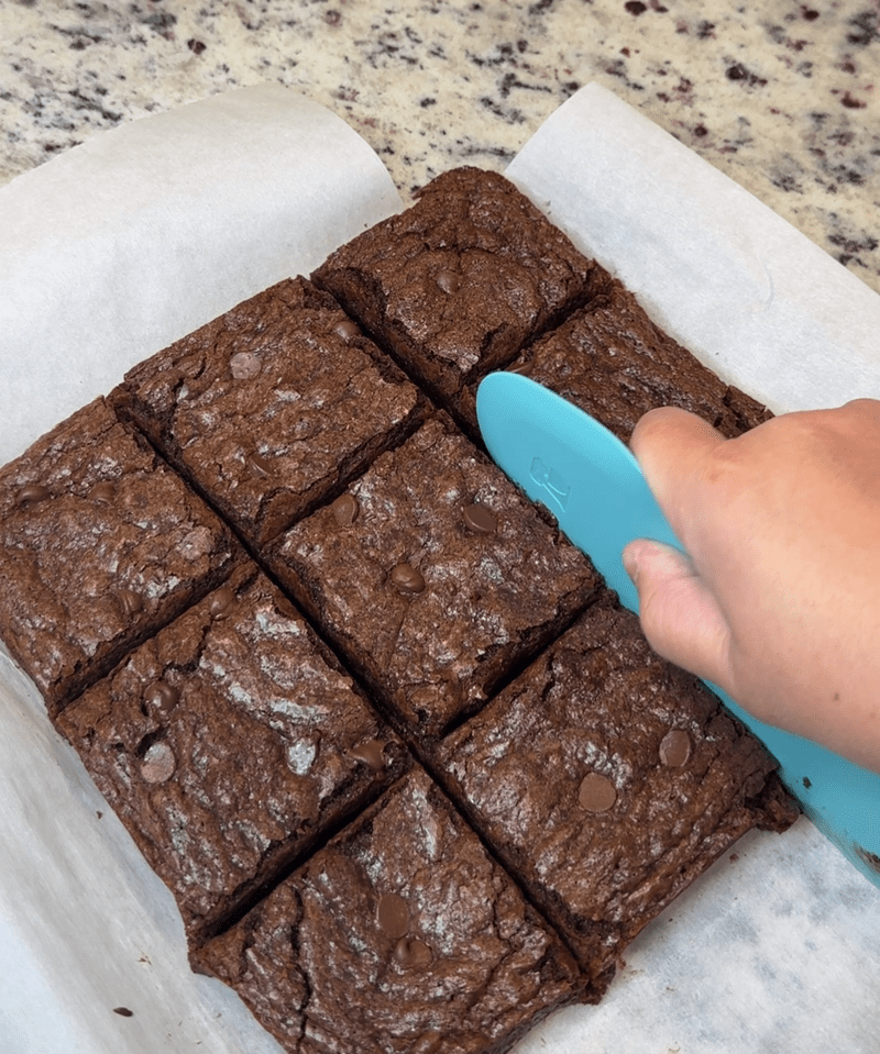 Cutting into brownies with a teal silicone bowl scraper. Brownies are mostly cut and shows brownies cut in a 3x3 grid into squares