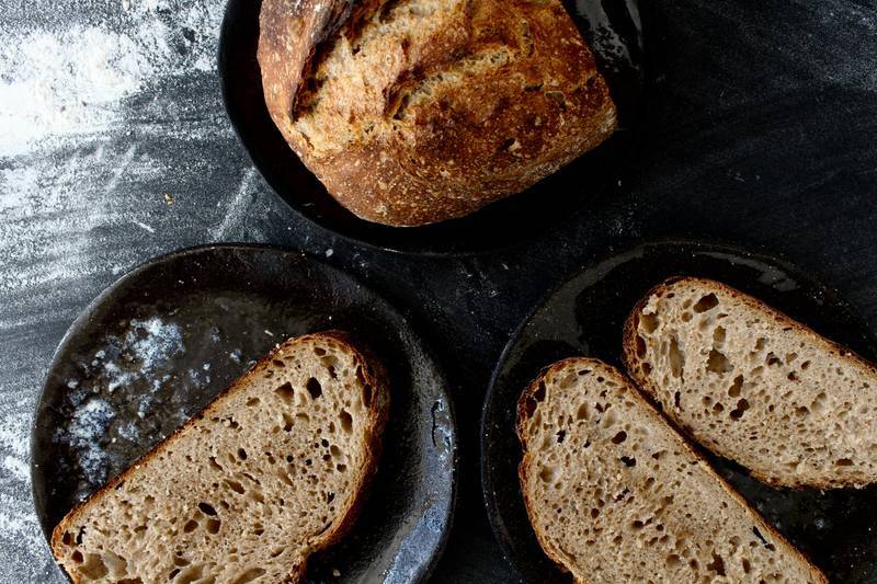 Freshly baked sourdough loaf and slices on dark plates with flour dusted surface.